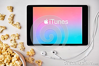 ITunes logo on the screen of the tablet laying on the white table and sprinkled popcorn on it. Apple earphones near the Editorial Stock Photo