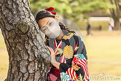 Woman in kimono wearing a mask leaned against the trunk of a pine tree. Stock Photo