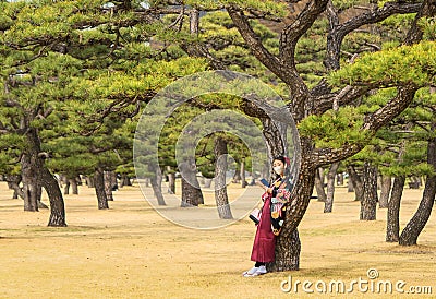 Woman in kimono wearing a mask leaned against the trunk of a pine tree. Stock Photo