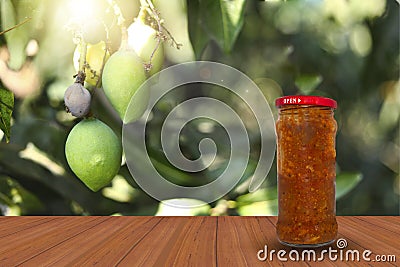 A jar of spicy mango pickle on a wooden table surface against the natural backdrop of a beautiful mango tree Stock Photo