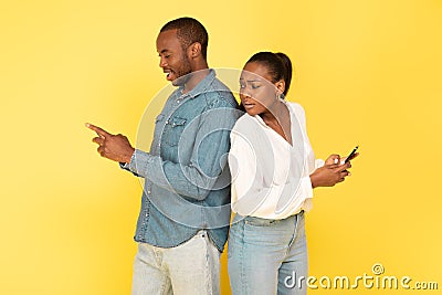 Jealous African American Wife Peeking At Husband's Smartphone, Yellow Background Stock Photo