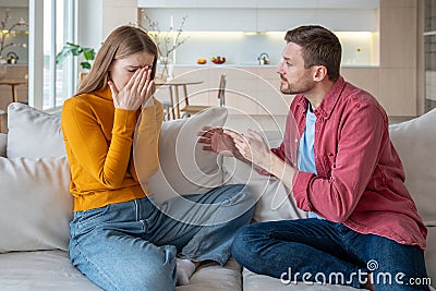 Jealous furious abused man talking screaming at crying woman wife sitting on couch in living room. Stock Photo