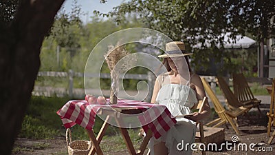 Jeune fille rurale sous chapeau de paille et robe blanche assise à la petite table avec vase à herbe décorative lisant le banque de vidéos Jeune fille rurale sous chapeau de paille et robe blanche assise à la petite table avec vase à herbe décorative lisant le banque de vidéos