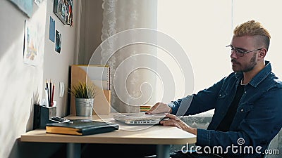 Jeune indépendant à la maison beau avec des verres et des cheveux jaunes fonctionnant à la maison, utilisant un ordinateur portab banque de vidéos Jeune indépendant à la maison beau avec des verres et des cheveux jaunes fonctionnant à la maison, utilisant un ordinateur portab banque de vidéos