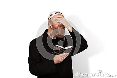 Jewish man holding Siddur covering eyes with hand on white background. Bearded Jew hasid with sidelocks in white kippah Stock Photo