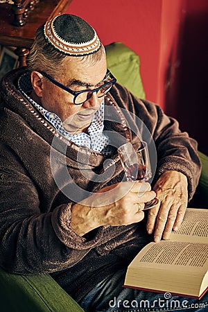 Jewish senior in the armchair reading a torah book and drinking kosher wine Stock Photo