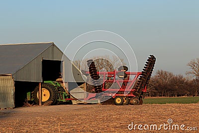 A John Deere tractor with a Sunflower oneway disc on the back with tree`s and blue sky with a barn. Editorial Stock Photo