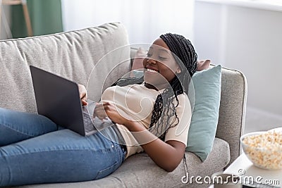 Joyful african lady laying on couch at home, using laptop Stock Photo