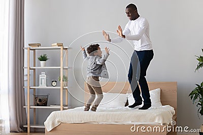 Joyful african american young man jumping on bed with son. Stock Photo