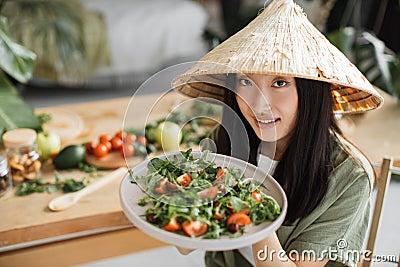 Joyful asian woman in traditional conical hat holding healthy salad from organic vegetables Stock Photo