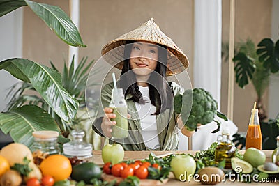 Joyful asian woman in traditional conical hat making smoothies using broccoli Stock Photo