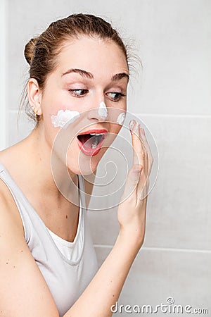 Joyful beautiful young woman with tied hair applying hydrating cream Stock Photo