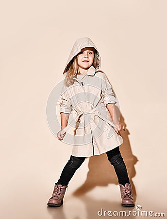 Joyful blonde kid girl tightens her belt on a beige coat happy smiling on beige Stock Photo