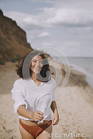 Joyful boho girl in white shirt running on sunny beach. Carefree stylish woman in swimsuit and shirt relaxing on seashore. Summer Stock Photo