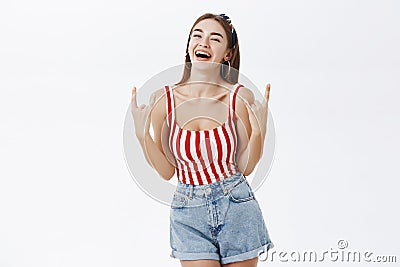 Joyful carefree stylish pinup girlfriend in striped top and denim shorts showing rock n roll gesture laughing amused Stock Photo