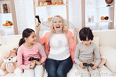 Joyful grandmother with cheerful grandchildren playing on game console sitting on couch. Stock Photo