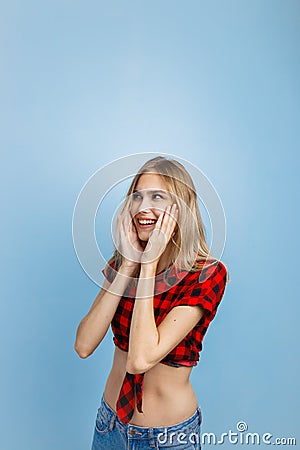 Joyful handsome young blond girl with blue eyes wearing red checkered shirt and blue jeans, with hands on her head Stock Photo