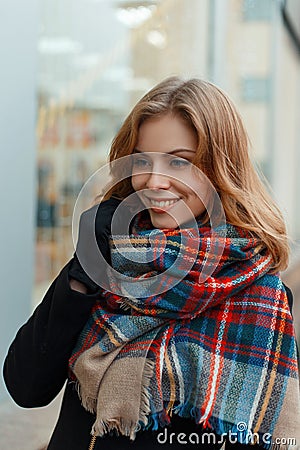 Joyful happy young woman in a fashionable warm wool scarf in a stylish black coat in black gloves is standing and smiling Stock Photo