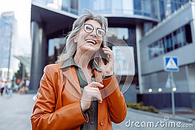 Joyful hoary haired senior lady talks on cellphone on modern city street Stock Photo