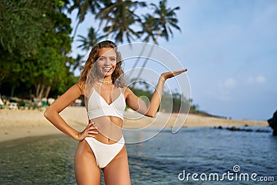 Happy woman in bikini presents invisible product on palm at tropical beach. Female model showcasing, advertising space Stock Photo