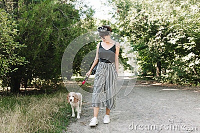 Joyful lady in white sneakers walking with beagle dog in park in sunny day, enjoying good weather. Outdoor full-length Stock Photo