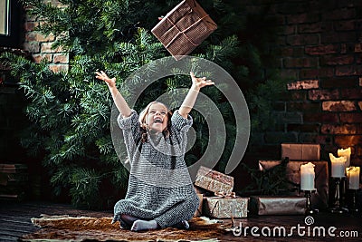 Joyful little girl with blonde curly hair wearing a warm sweater throws up a gift box while sitting on a floor next to Stock Photo