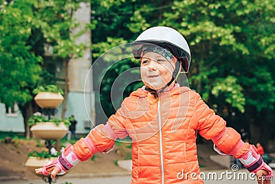 A joyful little girl in a helmet puts her hands to the sides. Beautiful child plays outdoors in the city park Stock Photo