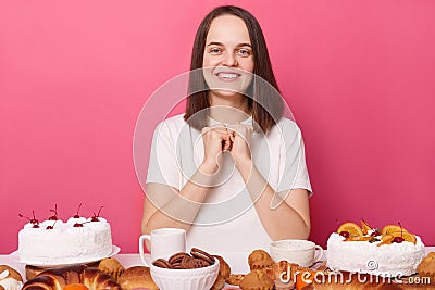 Joyful smiling delighted brown haired woman in white t shirt sitting at table with delicious desserts isolated over pink Stock Photo