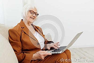 a joyful, sweet, gray-haired elderly woman in a brown suit is sitting on a beige sofa in a bright room working remotely Stock Photo