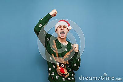 Joyful young man in santa hat and warm christmas sweater stands against a blue background and rejoices in victory. Happy man won, Stock Photo