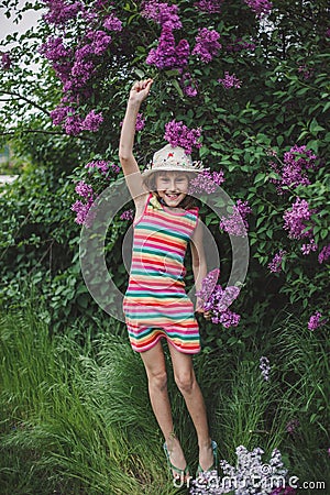 A jumping laughing girl of European appearance in a striped dress and a hat in a lilac garden Stock Photo