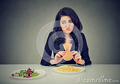 Junk food vs healthy food. Hesitant woman with cheeseburger looking at vegetables salad Stock Photo