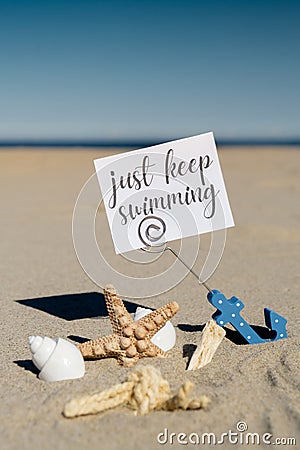 JUST KEEP SWIMMING text on paper greeting card in anchor paper holder and starfish seashell summer vacation decor. Sandy Stock Photo