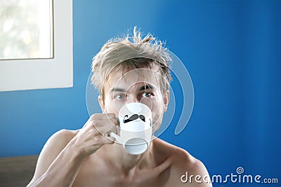 Just woke up and disheveled man drinking fresh coffee or tea from mug with painted black mustache, good weekend or weekday morning Stock Photo