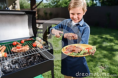 Kid boy in apron preparing tasty stakes on barbecue grill outdoors Stock Photo