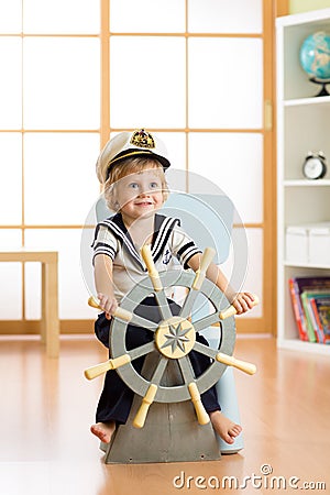 Kid boy dressed as a captain or sailor plays on chair like ship in his room. Child rotates the wooden steering wheel. Stock Photo