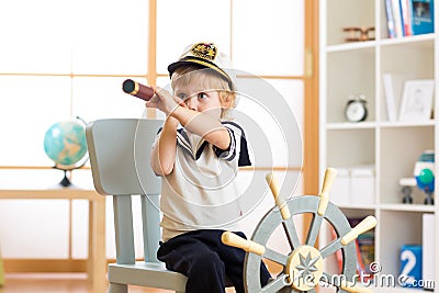 Kid boy dressed like a captain or sailor plays on chair as ship in his room. Child looks through telescope. Stock Photo