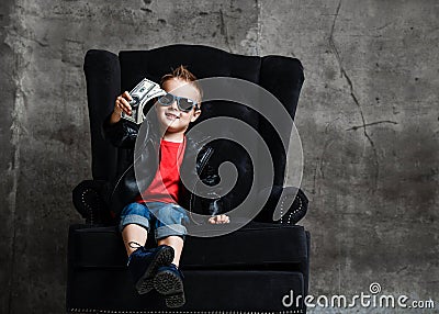 Kid boy millionaire in leather jacket and red t-shirt is sitting in big luxury armchair handing us a bundle of dollars cash Stock Photo