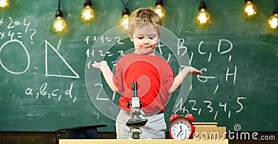 Kid boy near microscope, clock in classroom, chalkboard on background. First former confused with studying, learning Stock Photo