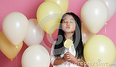 Kid girl with balloons in princess dress with tiara. Eyes closed. Holds yellow lollipop. Stock Photo