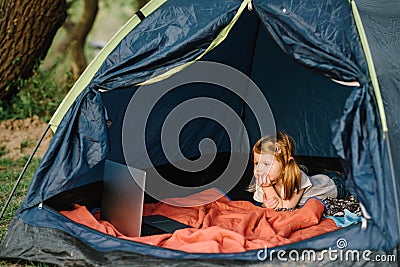 Kid girl in a campaign in a tent. Family summer vacation in nature. Children tourism. Child using laptop in the tent at the Stock Photo