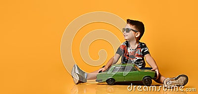 Kid in sunglasses, camouflage t-shirt, sneakers. Sitting on floor, holding green model of retro car, posing on orange background Stock Photo