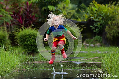 Kid playing out in the rain. Children with umbrella and rain boots play outdoors in heavy rain. Little boy jumping in muddy puddle Stock Photo