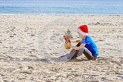 Kid in red Santa hats and pineapple as Christmas tree sitting at sea beach during Christmas vacation Stock Photo