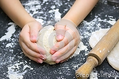 Kid`s hands, some flour, wheat dough and rolling pin on the black table. Children hands making the rye dough for backing bread. S Stock Photo