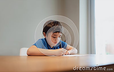 Kid siting on table doing homework,Child boy holding black pen writing on white paper,Young boy practicing English words at home. Stock Photo