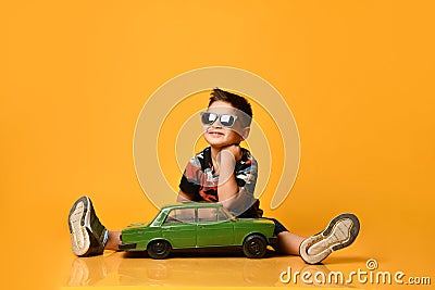 Kid in sunglasses, camouflage t-shirt, sneakers. Sitting on floor, holding green model of retro car, posing on orange background Stock Photo