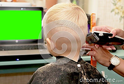 A little boy is cutting a hairdresser in the salon. The kid is watching a cartoon. Green screen on a laptop for signature Stock Photo