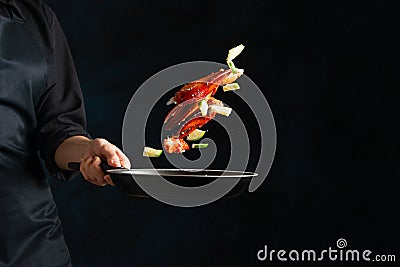 King crab legs and lime are flying in a frying pan on dark blue background. The professional chef in black uniform keeps pan with Stock Photo