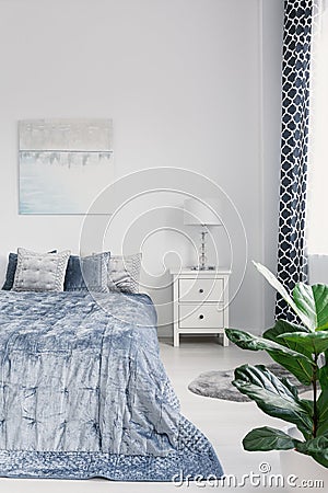 King size bed with elegant blue bedding, white nightstand with lamp and painting on the wall in luxury bedroom interior, real phot Stock Photo